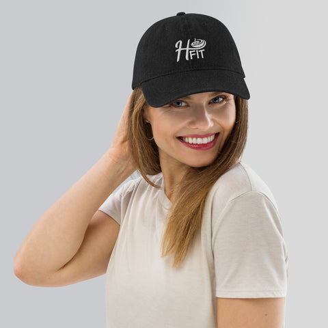 Smiling woman in a beige t-shirt and H-Fit Black Denim Hat with an adjustable strap, touching the back of her head, posing against a light gray background.