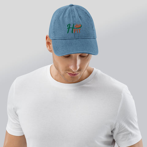 A man wearing the H-Fit Blue Denim Hat—pigment-dyed blue with HFIT embroidered in green and orange—pairs it with a plain white t-shirt, looking down against a light gray background. The 100% cotton cap has an adjustable strap for comfort.