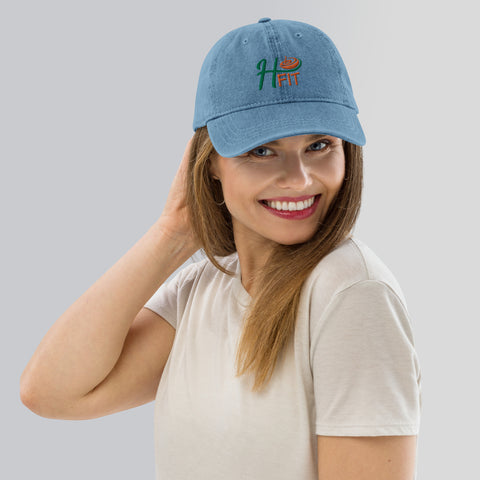 A smiling woman with straight light brown hair wears the H-Fit Blue Denim Hat and a light beige t-shirt, posing with her hand in her hair against a light gray background.
