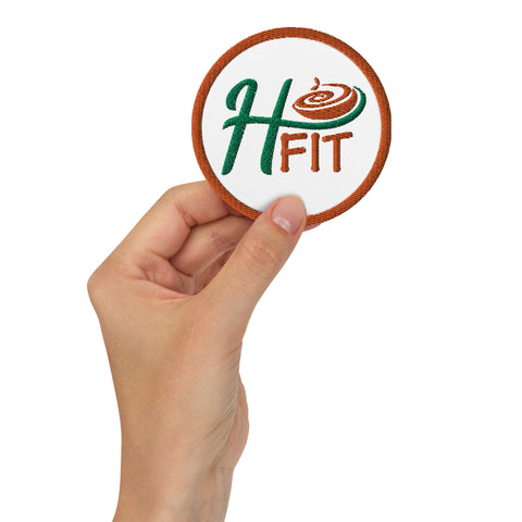 A hand holds an Embroidered patch with a brown border, featuring H FIT in green and orange thread and a swirl pattern inside the letter H. This round sew-on patch is part of our Embroidered patches collection.