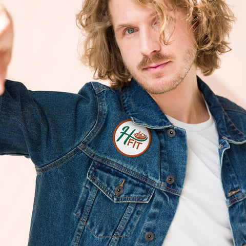 A man with wavy blond hair and a beard wears a denim jacket with an Embroidered patch—an H FIT circular pin featuring a bowl of soup. He faces the camera with a neutral expression against a light background.