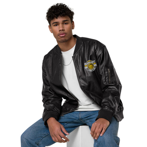 A young man with curly hair sits on a white block against a plain white background, wearing the Lions Club Leather Bomber Jacket with a patch on the left side, a white t-shirt, blue jeans, and a thin chain necklace.