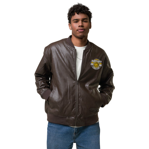 A young man stands with his hands in his pockets, wearing the Lions Club Leather Bomber Jacket in brown over a white T-shirt and blue jeans. The jacket features a yellow and white embroidered patch on the chest.