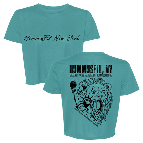The Turquoise Hummus Fit Script Crop Tee features HummusFit New York in black script on the front and a bold back graphic of a roaring lion with fork and dumbbell, plus HUMMUSFIT NY text and meal prep info on soft four-way stretch fabric.