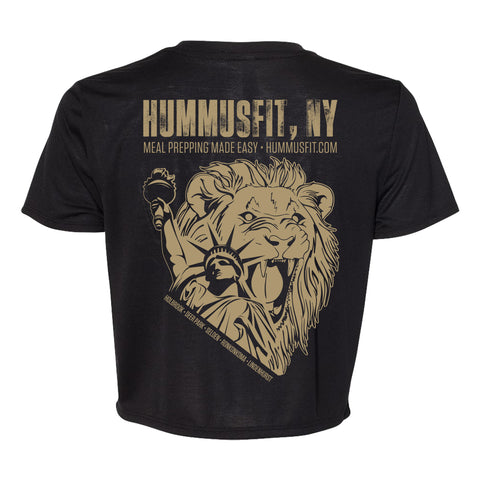 The Women’s Crop Tee is a trendy black shirt featuring a large tan graphic of a roaring lion and the Statue of Liberty, with “HUMMUSFIT,” “NY Meal Prepping Made Easy,” and “HUMMUSFIT.COM” printed above.