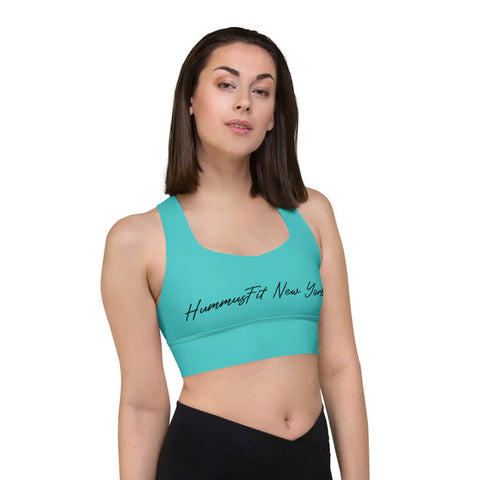 A woman in a turquoise Hummus New York Sports Bra made from compression fabric, paired with black leggings, stands against a plain white background.