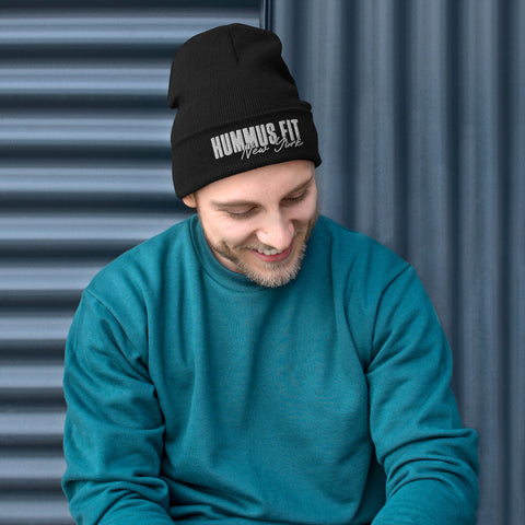 A man smiles and looks down while wearing a teal sweatshirt and the Hummus Fit NY Embroidered Beanie, sitting in front of a ribbed, dark blue metal wall.