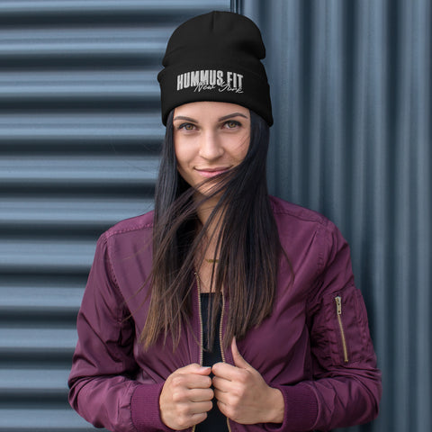 A woman in a maroon jacket and a Hummus Fit NY Embroidered Beanie stands before a corrugated metal wall, smiling slightly as the wind lifts her hair.