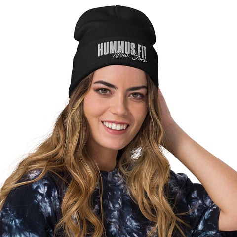 A smiling woman with long, wavy hair wears the Hummus Fit NY Embroidered Beanie and a dark tie-dye shirt, holding one hand to her head against a plain white background.