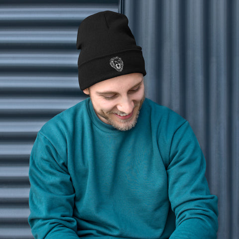 A man wearing the Unisex Embroidered Beanie and a teal cotton blend sweatshirt sits in front of a dark corrugated metal wall, looking down and smiling.