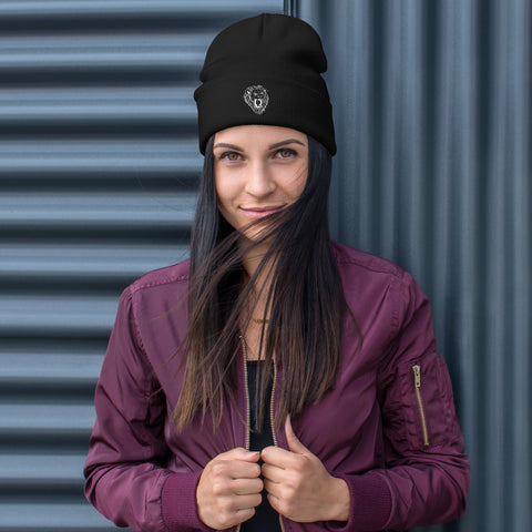 A woman with long dark hair wears a Unisex Embroidered Beanie and a maroon jacket, standing before a corrugated metal wall, holding her jacket lapels and smiling slightly.