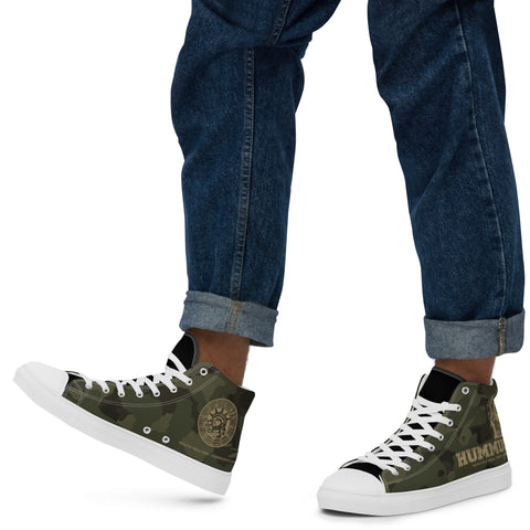 A man walks on a white background wearing rolled-up blue jeans and Men’s Army Camo High Top Canvas Shoes—handmade with a camouflage print, white soles and laces, plus text and a circular side design.