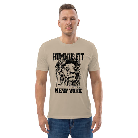 A man wears the Unisex Hummus Fit New York Desert Dust T-Shirt featuring a black lion face graphic and HUMMUSFIT NEW YORK text, standing facing forward against a plain white background.