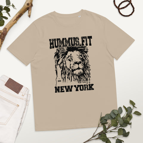 Unisex Hummus Fit New York Desert Dust T-Shirt made from organic cotton, featuring a black lion face graphic, New York skyline, and “HUMMUS FIT NEW YORK.” Displayed on a flat surface with jeans, beads, twigs, and green leaves.