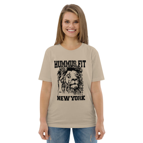 A smiling woman with straight light brown hair wears the Unisex Hummus Fit New York Desert Dust T-Shirt, beige with a black lion graphic and HUMMUS FIT NEW YORK text, standing against a plain white background.