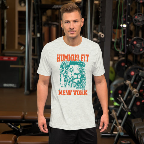 A man stands in a gym wearing the Unisex Hummus Fit New York Ash T-Shirt, made from 100% cotton and featuring a stylized lion graphic, with gym equipment and weights visible in the background.