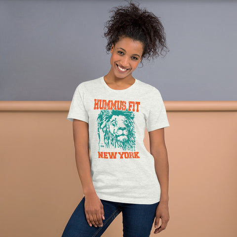 A smiling woman with curly hair wears the Unisex Hummus Fit New York Ash T-Shirt, 100% cotton in light gray, featuring an illustrated lion and bold orange and teal text. She stands against a beige and gray background.