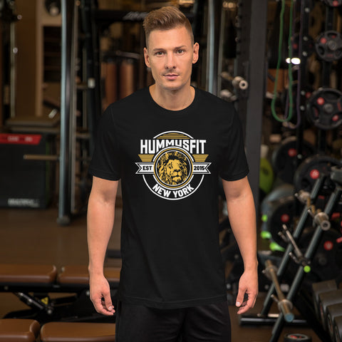 A man with short blonde hair stands in a gym, wearing a comfortable Unisex Gold Hummus Fit T-Shirt featuring a lion’s head graphic. Weightlifting equipment and gym machines are visible in the background.