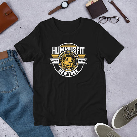 The Unisex Gold Hummus Fit T-Shirt, made from soft cotton and featuring a bold lions head logo with EST 2015, is styled on concrete alongside jeans, shoes, glasses, a watch, and accessories.