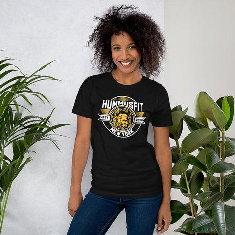 A smiling person with curly hair wears a Unisex Gold Hummus Fit T-Shirt, standing between two green potted plants against a light-colored wall.