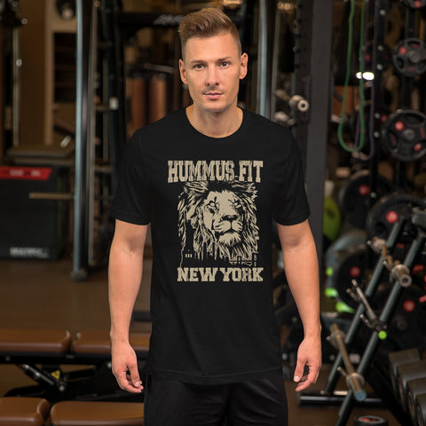A man with short blond hair stands in a gym wearing the Unisex Hummus Fit New York Black T-Shirt, which features a lion’s face and text. Gym equipment and weights are visible in the background.