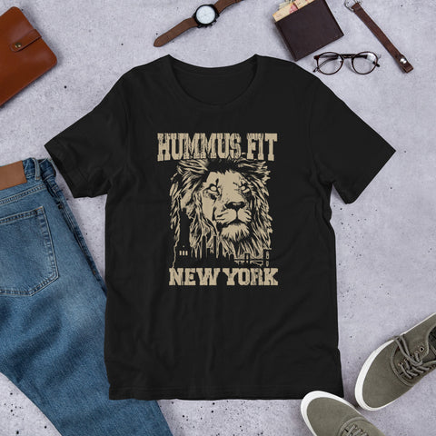 The Unisex Hummus Fit New York Black T-Shirt is a lightweight, 100% cotton tee with a lion’s face and bold logo, shown laid flat on a gray surface alongside jeans, shoes, glasses, a watch, and a brown wallet.