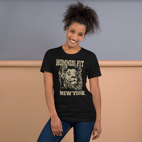 A smiling woman with curly hair wears the Unisex Hummus Fit New York Black T-Shirt, featuring a lion graphic, standing against a two-toned beige and blue-gray background.