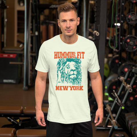 A man stands in a gym wearing the Unisex Hummus Fit New York Citron T-Shirt, featuring a graphic lion and text. Gym equipment, weights, and benches appear in the background.