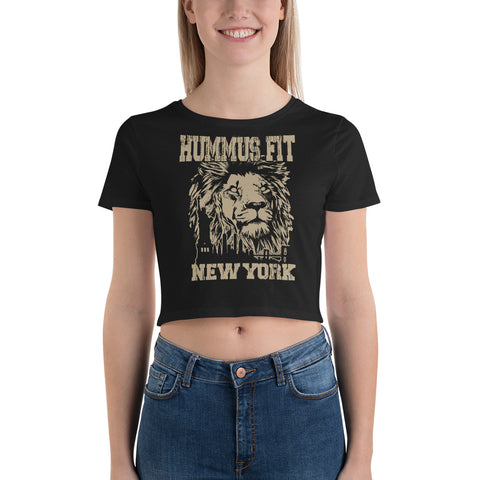 A smiling woman models the Women’s Crop Tee, featuring a bold lion graphic and “HUMMUS FIT NEW YORK” text in distressed letters. This slim fit crop top stands out against a plain white background.