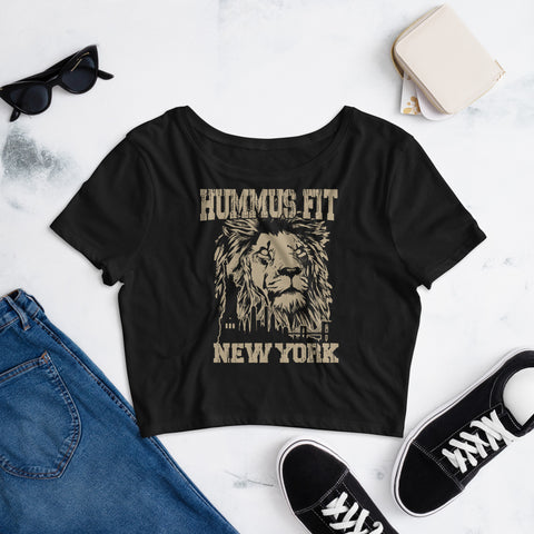 The Women’s Crop Tee is a trendy black top with a lion face graphic and “HUMMUS FIT NEW YORK,” shown next to sunglasses, a beige wallet, blue jeans, and black-and-white sneakers on a white surface.