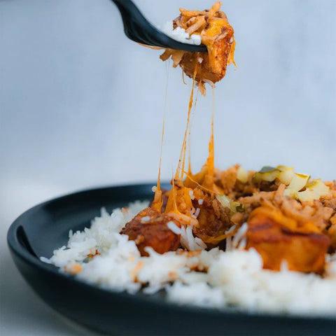 A black spoon lifts cheesy, saucy meat and vegetables above a plate of white rice on a black dish, with melted cheese stretching between the spoon and the food.