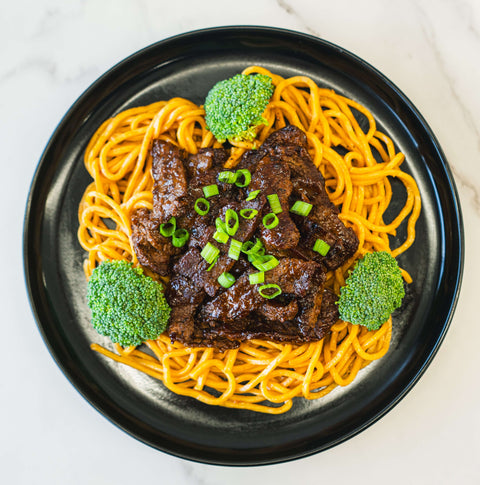 A black plate with yellow noodles topped with slices of glazed beef, garnished with chopped green onions and three pieces of broccoli on the side. The plate is on a white marble surface.