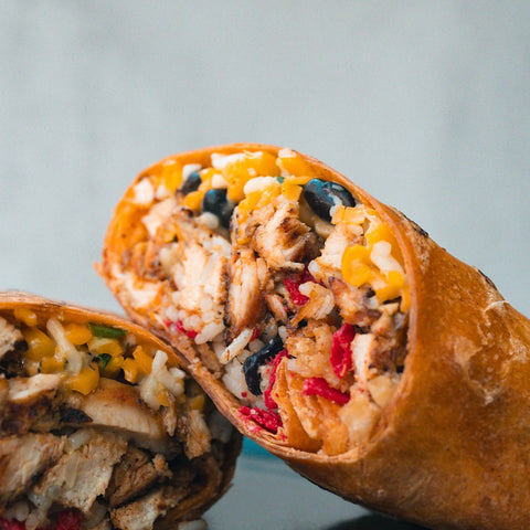 A close-up of a burrito cut in half, showing grilled chicken, rice, black beans, cheese, corn, and red peppers inside a toasted tortilla.