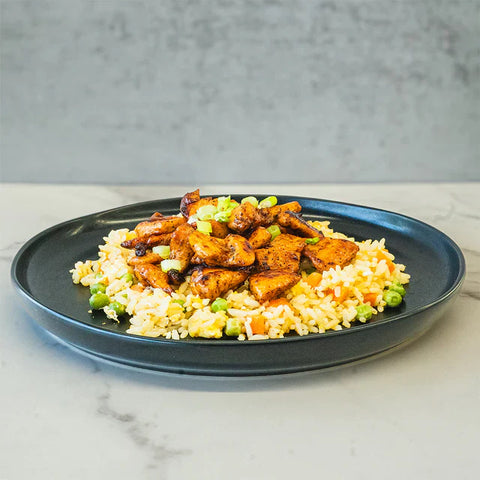 A black plate of fried rice with peas, carrots, and chunks of grilled chicken, set on a marble surface with a gray background.