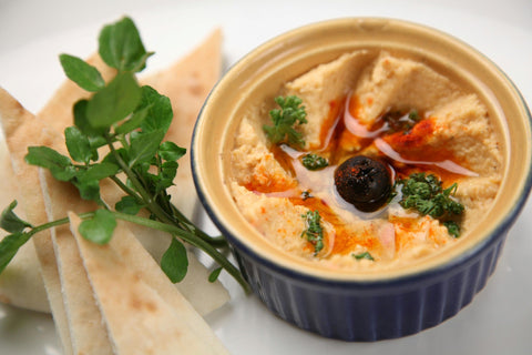 A bowl of hummus garnished with parsley, olive oil, paprika, and a black olive, served with pita bread slices and fresh greens on the side.