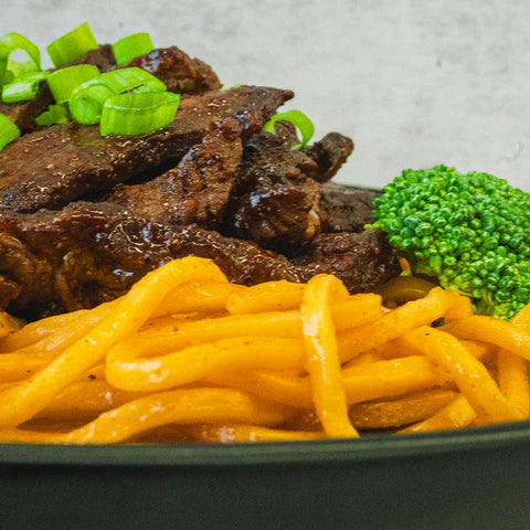 A close-up of a dish with glazed beef topped with chopped green onions, served alongside yellow noodles and a piece of broccoli.