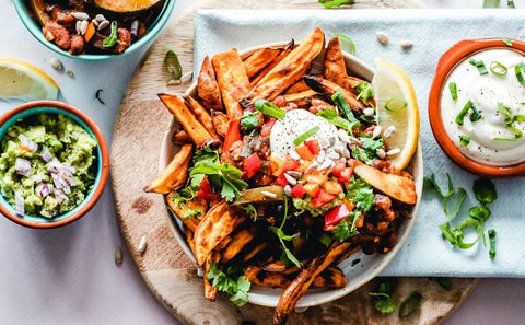 A plate of sweet potato fries topped with sour cream, chopped tomatoes, beans, cilantro, and seeds, served with guacamole and dip on a wooden board.
