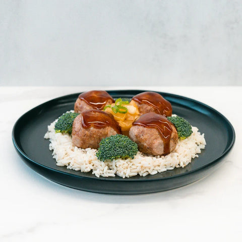 A black plate with rice topped with meatballs and brown sauce, surrounded by broccoli florets and garnished with green onions on a white background.