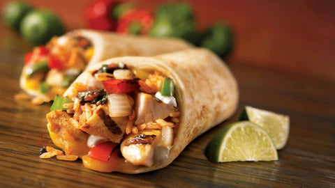 Two burritos filled with grilled chicken, rice, bell peppers, onions, and sauce sit on a wooden surface, with lime wedges and blurred vegetables in the background.