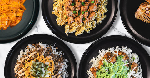 Five black plates with different meals, including pasta with chicken, rice with seasoned meat, rice with green sauce and chicken, nachos with cheese, and a pastry, arranged on a marble surface.