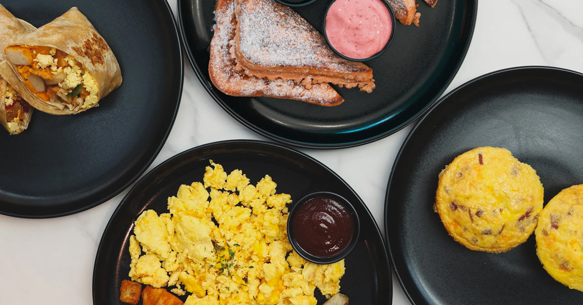 Flat lay of a black plate breakfast spread with a halved burrito, chocolate-drizzled French toast with pistachios and green dip, scrambled eggs, sausage, hash browns with ketchup, and two bacon egg muffins on a white marble surface.