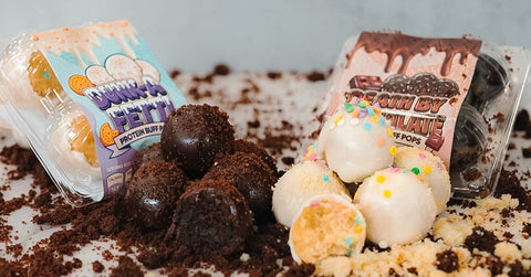 Two packs of cake pops sit on a surface covered with cake crumbs. One pack is chocolate with frosting, the other is white with colorful sprinkles. Several cake pops are outside their packaging.