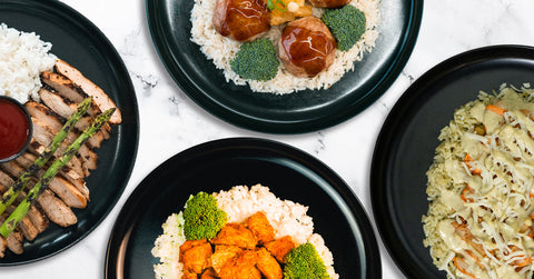 A black plate with seasoned grilled chicken pieces served on a bed of fried rice with peas and carrots, placed on a marble surface with a gray background.