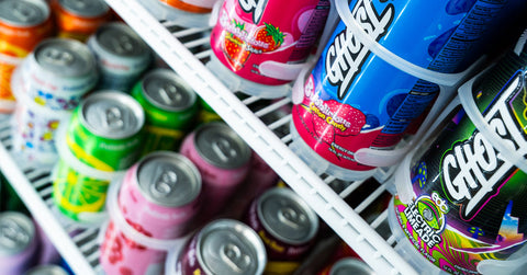 Rows of colorful canned and bottled soft drinks are stacked neatly on shelves inside a refrigerator, featuring various bright labels and flavors.