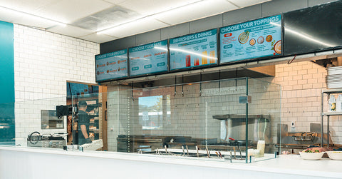 A modern fast-casual restaurant counter with a glass barrier, digital menu boards overhead, white tile walls, and a staff member preparing food behind the counter.