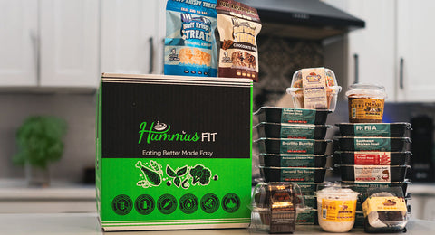 A green Hummus Fit meal box sits on a kitchen counter surrounded by stacks of prepackaged healthy meals, snacks, and treats, with a blurred kitchen background.
