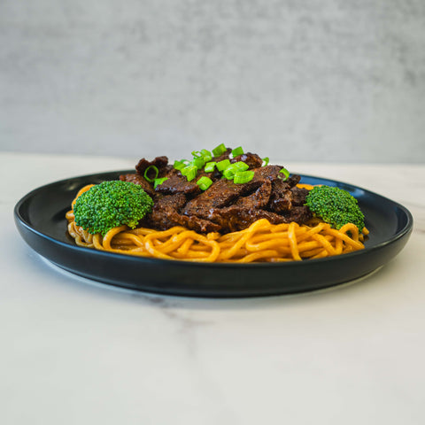 A black plate with noodles topped with sliced beef and chopped green onions, garnished with two pieces of broccoli, set on a white surface with a gray background.