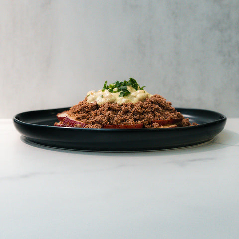 A black plate featuring a mound of crumbled brown food topped with a creamy yellow sauce and chopped green herbs, set against a light gray background.