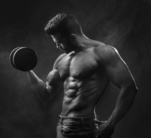 A muscular man in jeans poses shirtless while lifting a dumbbell in one hand, highlighting his bicep and defined physique against a dark, smoky background.