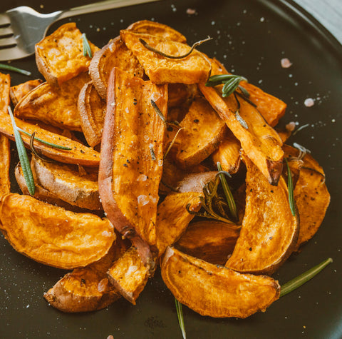 A plate of roasted sweet potato wedges garnished with rosemary and sprinkled with coarse salt, served on a dark dish with a fork in the background.
